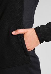 Zwarte fleecjas met een gestructureerd, gewatteerd patroon op het zakgedeelte; hand in een zijzak gestoken, met zichtbare ritsdetails.