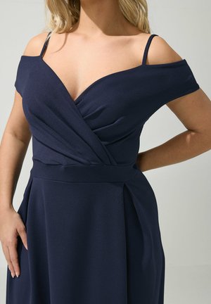 Woman wearing a navy blue off-shoulder dress with thin straps, a wrap-style bodice, and a fitted waistband.