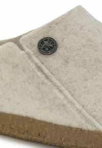 Light gray felt slipper with a brown rubber sole. Features a circular black logo accent. Texture appears soft and durable.