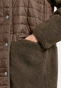 Gekwalte bruine buitenlaag met een zachte, gestructureerde bruine fleece voering en zak. Kenmerkt zich door drukknoopsluiting en een contrasterende rand.