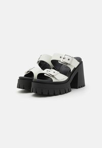 Sandale cu toc gros, cu o talpă neagră groasă, curele din piele albă și catarame din argint. Design deschis la vârf, cu o platformă texturată.