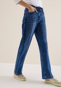 Vaqueros azules de corte relajado con cintura ajustable, cinco bolsillos y una textura de denim suave. Combinados con zapatillas de color claro.