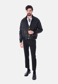 Giacca bomber in pelle nera, cardigan verde bottonato sotto, camicia bianca, pantaloni neri e scarpe nere con dettagli a fibbia.