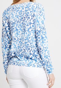 Blaues Langarmoberteil mit einem runden Muster in unterschiedlichen Größen, das eine weiche Textur aufweist. Heller weißer Grund mit Akzenten in Hellblau und Dunkelblau.