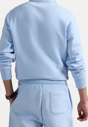 Person trägt hellblauen Langarm-Sweatshirt und passende Hose mit Gesäßtasche, Armbanduhr am linken Handgelenk, steht vor weißem Hintergrund.