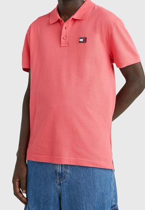 Mann trägt ein korallenrosa kurzärmliges Polohemd mit Tommy Jeans-Logo und blaue Jeansshorts vor einfachem Hintergrund.