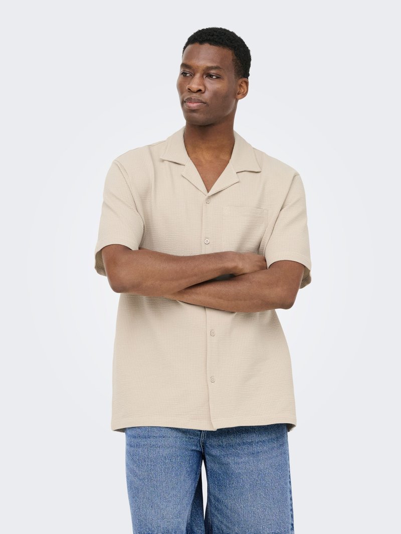 Homme les bras croisés portant une chemise beige à manches courtes boutonnée et un jean bleu, regardant légèrement sur le côté devant un fond uni.