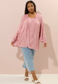Blusa a maniche lunghe rosa chiaro con tessuto testurizzato, bottone frontale, scollatura a volant e dettagli plissettati; abbinata a jeans cropped blu chiaro.
