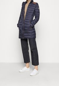 Marineblauer Puffermantel mit einem Reißverschluss und Druckknopfverschluss, braunen Schulterakzenten, zwei Seitentaschen und einem hohen Kragen, getragen über dunklen Jeans und weißen Sneakers.