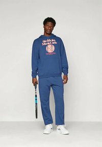 Tamsiai mėlynas džemperis su grafiniu spaudiniu ir atitinkamomis sportinėmis kelnėmis. Pagaminta iš medvilnės, laisvas pasiuvimas, dėvima su baltais sportbačiais ir laikant teniso raketę.