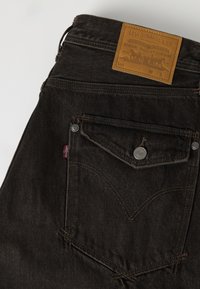 Πίσω μέρος μαύρου τζιν Levi's με ραμμένη καπάκι τσέπης με κουμπί, κόκκινη ετικέτα Levi's και καφέ δερμάτινη ετικέτα μάρκας στη μέση.