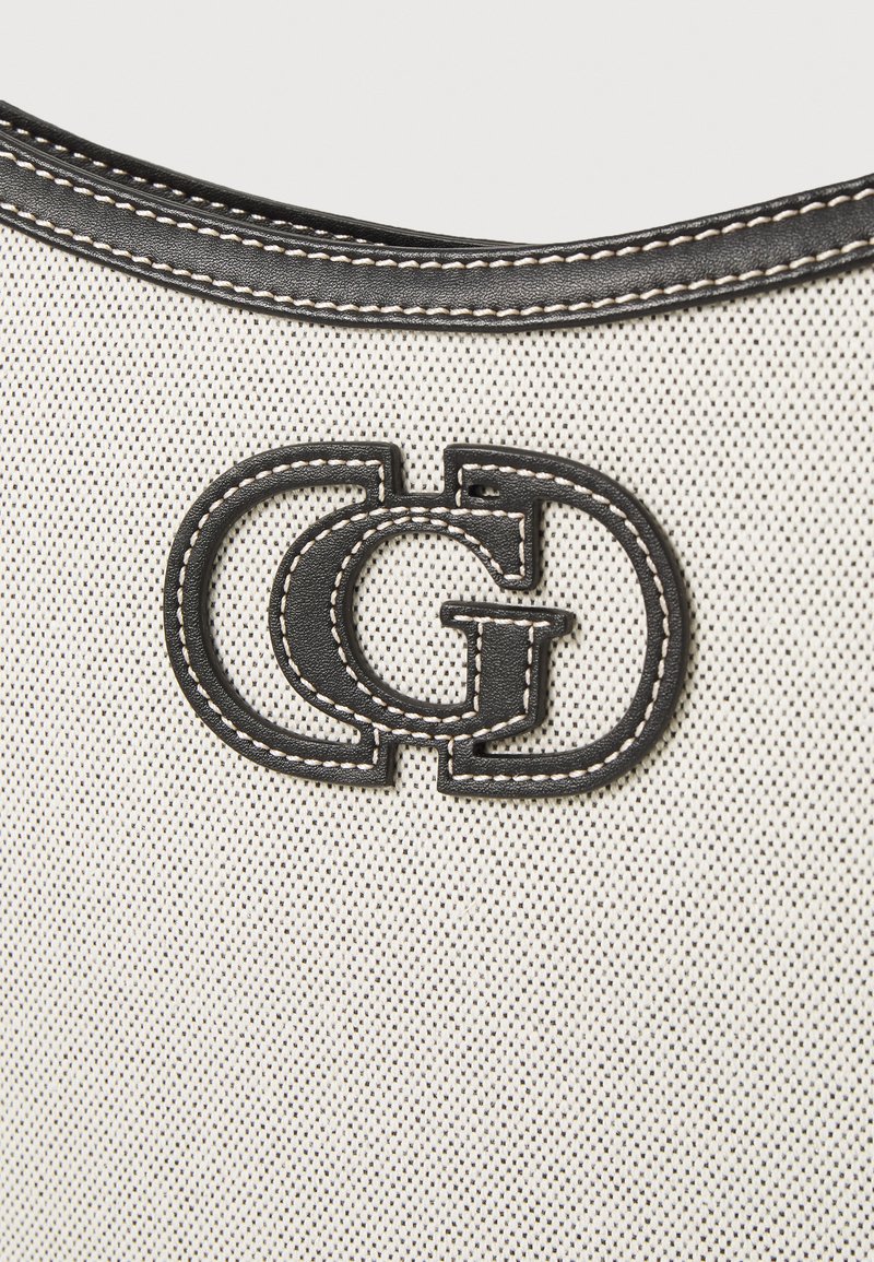 Canvas tas van crème stof, met een prominent zwart leren logo en contrasterende witte stiksels. Gladde textuur, afgeronde randen.