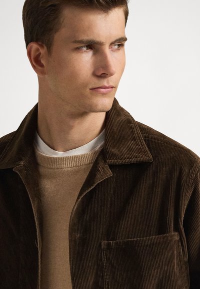 Καφέ μπουφάν από κορδόνι με γιακά σε σχήμα V, τσέπη στο στήθος και υφή υφάσματος, φορεμένο πάνω από μπεζ πουλόβερ και λευκό πουκάμισο.