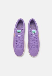 Fioletowe zamszowe sneakersy z płaskimi sznurówkami, białą gumową podeszwą i zieloną metką z logo na języku. Teksturowane wewnętrzne wykończenie z geometrycznym wzorem.