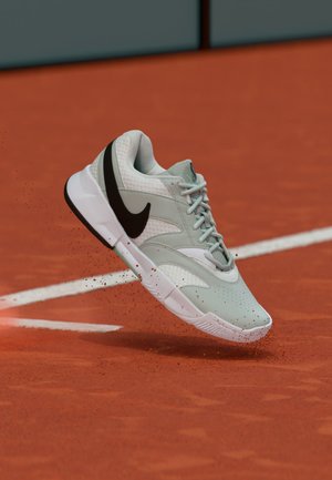 Zapatilla Nike gris claro y blanca con el símbolo Swoosh negro, suspendida en el aire sobre una pista de tenis de tierra batida roja, levantando polvo rojo.