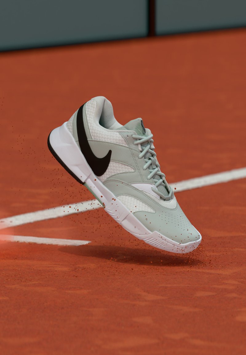 Jasnoszary i biały sneaker Nike z czarnym logo, zawieszony w powietrzu nad czerwoną, glinianą kortem tenisowym, unoszący czerwony pył.