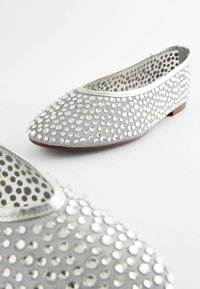 Sølvflade sko med en afrundet tå, prydet med cirkulære metalliske nitter, med en tekstureret overflade og tynd kanttrim.