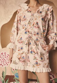 Rampers z bawełny w miękkim różowym kolorze z kwiatowym wzorem, ozdobiony falbankowym kołnierzem z koronki, elastycznym pasem i kieszeniami. Zawiera zabawne ilustracje i akcenty.