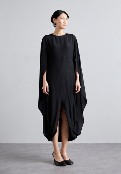 Vestido negro con un diseño fluido, mangas murciélago y una abertura frontal. Textura suave, hasta la rodilla con un dobladillo fruncido, combinado con tacones negros.