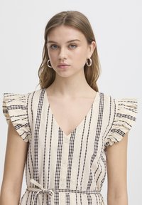 Abito beige con strisce verticali nere, maniche a volant, scollatura a V e chiusura con bottoni. Presenta una vita legata per una vestibilità regolabile.