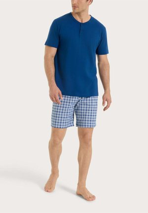 Uomo che indossa una maglietta henley a maniche corte blu e pantaloncini a quadretti blu e bianchi, in piedi a piedi nudi su uno sfondo semplice.