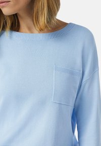 Hellblauer Pullover mit weitem Ausschnitt, gerippten Akzenten und einer kleinen aufgenähten Tasche auf der linken Seite. Weiche Textur mit lockerer Passform.