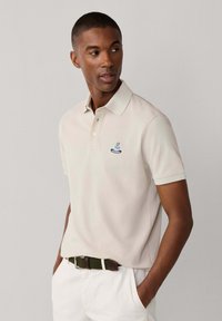 Mann trägt beige Polohemd mit kleinem blauem Logo auf der Brust und weiße Hose mit grünem gewebtem Gürtel, Hände in den Taschen, schaut nach rechts.