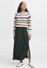 Pull en tricot rayé crème, vert et rose ; associé à une jupe maxi unie verte foncé avec une fente sur le côté et des baskets blanches.
