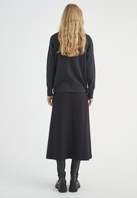 Černý svetr s dlouhými rukávy v kombinaci s černou sukní po kotníky. Modelka má na sobě černé kozačky do půli stehen. Měkká, strukturovaná tkanina.