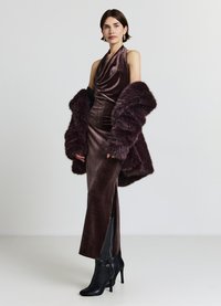 Abito halter in velluto bordeaux con scollo drappeggiato, spacco laterale, abbinato a un cappotto peloso in pelliccia bordeaux e stivali neri con tacco alto.