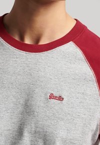 Närbild av en grå och röd raglantröja med en liten röd broderad "SuperDry"-logotyp på bröstet.