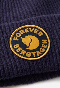 Dark purple ribbed knit fabric with a round yellow and black patch showing a fox head and text "FOREVER BERGTÅGEN".