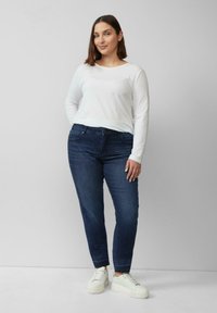 Weißes Langarmhemd, dunkelblaue Jeans und weiße Sneakers mit erhöhter Sohle, getragen von einem Model, das vor einem schlichten Hintergrund steht.