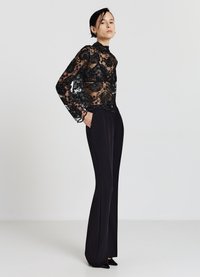 Blusa di pizzo nero con ricami floreali, maniche lunghe e colletto alto, abbinata a pantaloni neri su misura e scarpe nere a punta.