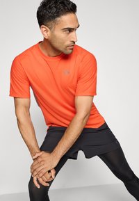 Mand iført en orange kortærmet skjorte, sorte shorts og sorte leggings, strækker benene, mens han kigger til siden mod en ensfarvet baggrund.