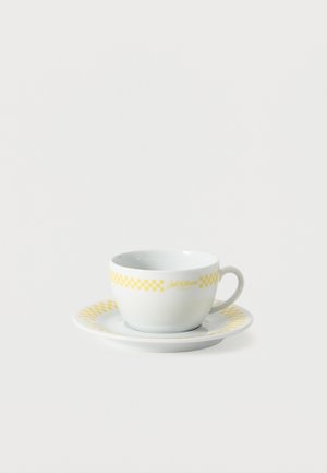 Tasse à thé en céramique blanche avec motif à carreaux jaune et soucoupe assortie sur un fond blanc uni.