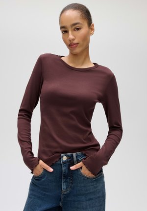Donna con capelli legati indossa una camicia aderente marrone scuro a maniche lunghe e jeans blu in denim con le mani nelle tasche.