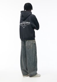Czarna bluza z kapturem z białym napisem na plecach, zestawiona z luźnymi szarymi jeansami. Napis zawiera szczegóły dotyczące marki i logo.