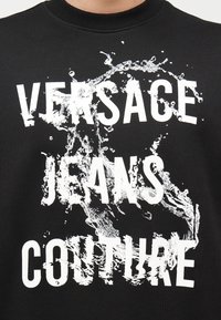 Μαύρο ύφασμα με λευκό κείμενο "VERSACE JEANS COUTURE", που καλύπτεται εν μέρει από λευκή σχεδίαση που μοιάζει με πιτσίλισμα νερού.