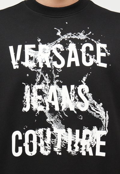 Tissu noir avec texte blanc "VERSACE JEANS COUTURE" partiellement dissimulé par un motif de splash blanc ressemblant à de l'eau.