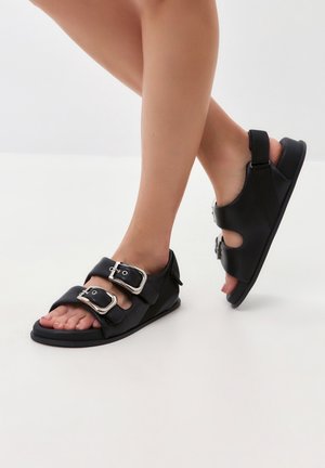 Musta sandaalit, joissa on kaksi leveää hihnaa hopeisilla soljilla, litteä pohja ja sivuilla leikatut aukot hengittävyyttä varten.