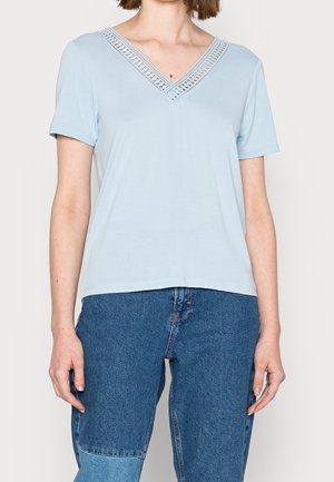 Vrouw draagt een lichtblauw T-shirt met korte mouwen, een V-hals met kanten rand en twee tonen blauwe spijkerbroek, staat met ontspannen armen.