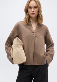 Νεαρή γυναίκα με ξανθά μαλλιά μέχρι τους ώμους, φορώντας καφέ μπλούζα με V λαιμόκοψη και μακριά μανίκια και μαύρο παντελόνι, κρατώντας μια μπεζ τσάντα στο πλάι της.