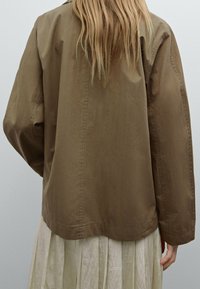 Braune, übergroße Jacke aus einem glatten, leichten Stoff. Mit abgesenkten Schultern und geradem Saum, getragen über einem leichten, plissierten Kleid.