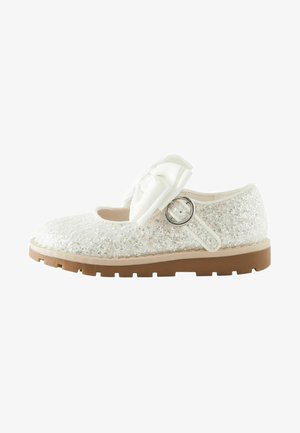 Chaussure blanche recouverte de paillettes, avec un bout arrondi, un grand nœud décoratif et un détail en anneau argenté sur la sangle. Semelle en caoutchouc beige.
