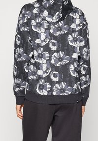 Person in einem schwarzen Kapuzenpullover mit großen weißen und grauen Blumenmustern und schwarzen Hosen, von hinten gezeigt.