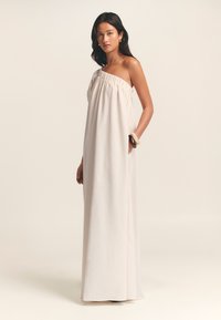 Abito maxi senza spalline in tessuto beige chiaro con una texture leggera, caratterizzato da un collo arricciato e tasche laterali per una funzionalità aggiuntiva.