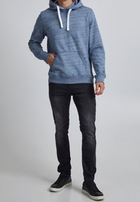 Sudadera con capucha azul y cordones blancos, bolsillo canguro, puños y dobladillo acanalados, combinada con jeans negros de corte ajustado y zapatillas negras.
