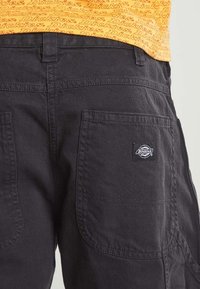 Black workwear pants with large back pockets, one featuring a small Dickies brand patch, worn with an orange shirt with repeated Dickies logo.