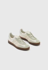 Pár béžových sneakers Adidas Gazelle se světle zelenými akcenty a gumovými podrážkami na bílém pozadí.
